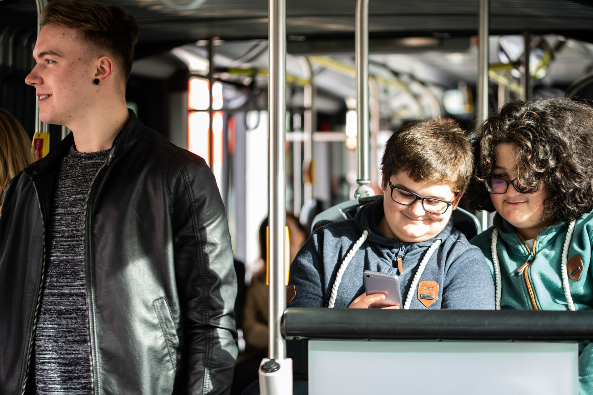 Due ragazzi seduti in autobus controllano insieme uno smartphone. Sulla sinistra un altro ragazzo in piedi che gli volta le spalle.